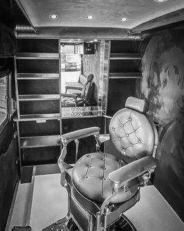 Black and white interior shot of a mobile barber van, focusing on a leather barber chair in the foreground and a mirrored reflection of the rear cabin, including shelving and a bench seat.