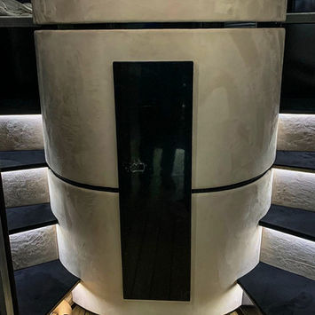 A futuristic cylindrical enclosure inside a luxury motorhome, positioned between two staircases with built-in strip lighting. The curved structure is finished in a smooth cream tone with a vertical black gloss panel at the center. The floor is lined with natural wood-look vinyl. The design hints at a bespoke wet room or central feature area, part of a sleek, modern RV interior layout.