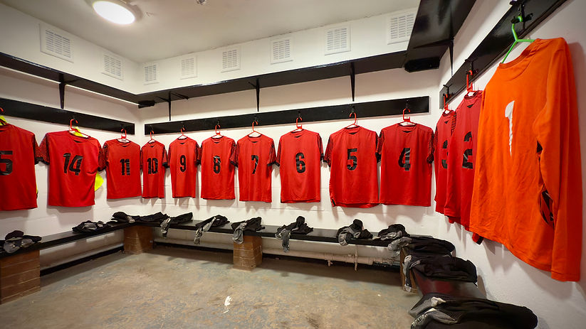 A football locker room with red and black team shirts neatly hung on wall hooks, numbered 1 through 14. The number 1 shirt is orange, indicating the goalkeeper's jersey. Beneath the shirts, benches hold black socks and folded kits, ready for players. The clean, organised setup suggests preparation for an upcoming match.
