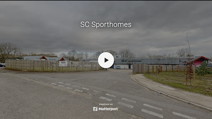 The launch screen of a Matterport virtual tour for SC Sporthomes, showing the exterior of the company’s main facility on an overcast day. The site features a large industrial-style building with corrugated metal cladding, branded with “SC Sporthomes” in bold black lettering. A wooden fence encloses part of the property, with a sign near the entrance gate indicating company and visitor access information. A blue car is parked inside the open gate, and landscaped areas with young trees and a small evergreen line the curved entrance drive. The Matterport interface includes a central play button and branding footer, inviting viewers to explore the immersive 3D experience of the SC Sporthomes business premises.