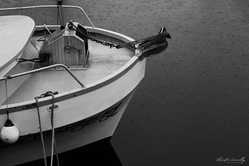 Bow of a boat with an anchor, ropes, and a buoy on a rainy day. Water shows rain ripples. Image is black and white, creating a calm mood.