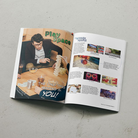 An image of a magazine open on a flat surface. The left page shows an ad for a fictional board game cafe called "Playspace" and the right page is a table of contents.