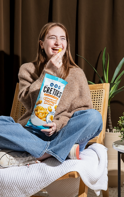 Young woman cross legged on a chair holding bag of Cretors caramel puffs and smiling.