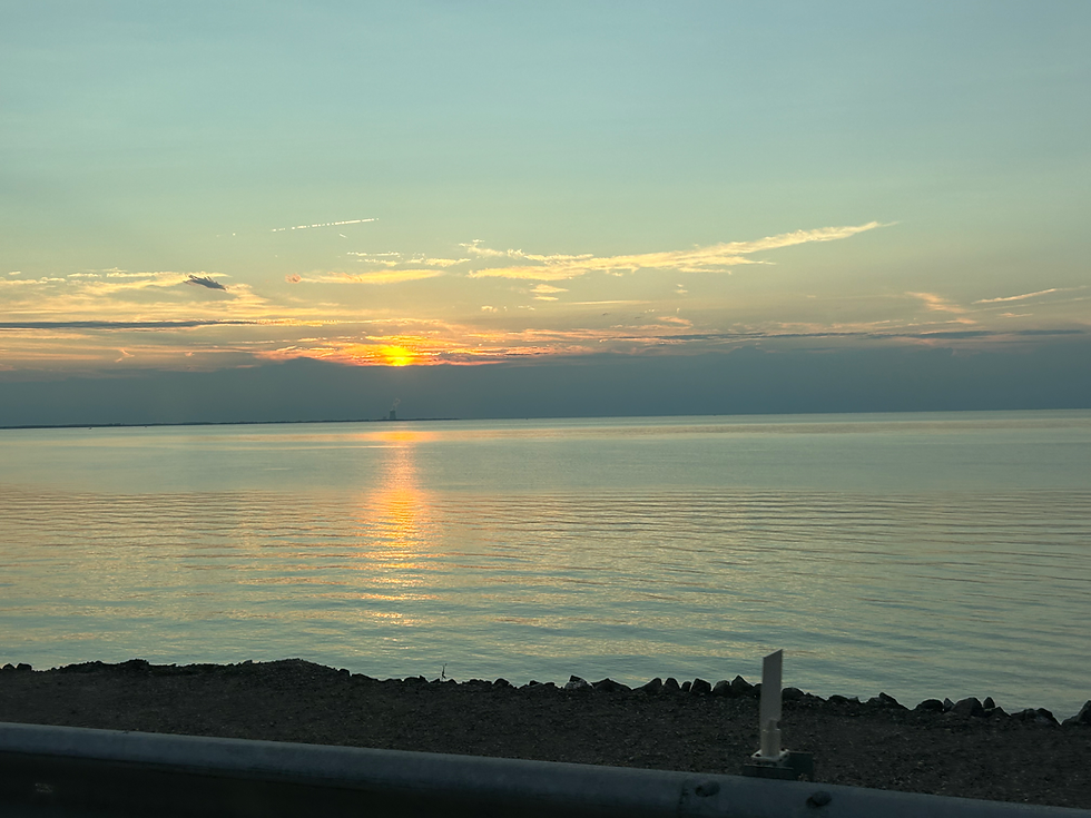 Golden sunset over calm water, orange reflection on the sea, with rocky shoreline in the foreground.