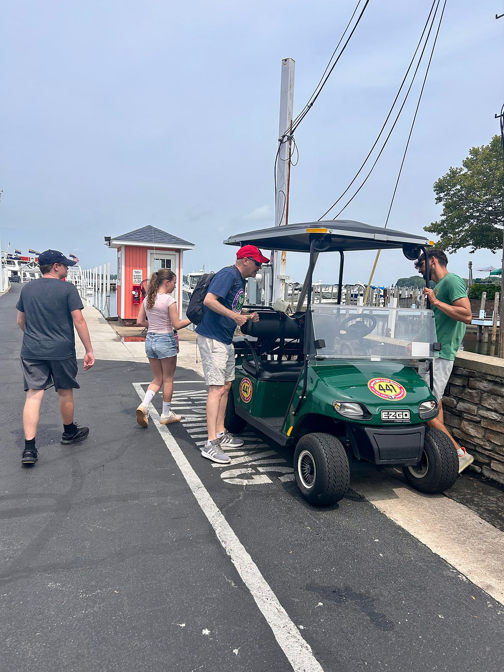 Four people gather around a green EZGO golf cart at a marina dock, with boats, posts, and a small red shed in the background.