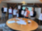 Group of people smiling and holding drawings in a room with wooden beams, a round table with paper and pens in the foreground.