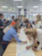 People draw on a long white paper at a table in a bright office, using colorful markers. The mood is collaborative and creative.
