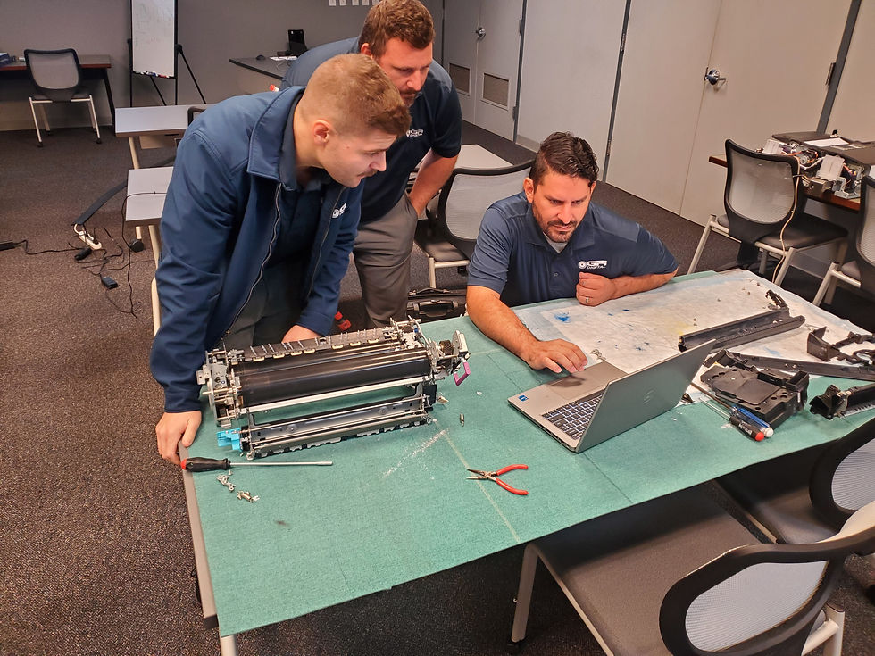 Three men in a workshop examine a printer assembly. One uses a laptop, while tools and parts are scattered on the green table. Focused mood.