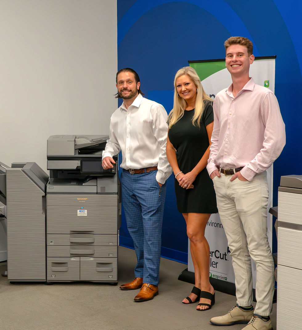 Three GFI Digital employees smiling beside a printer in an office with a blue accent wall. They wear business casual clothing. A poster is partially visible.
