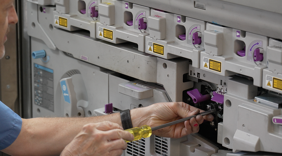 Technician using a screwdriver to repair a large, gray production machine with purple knobs.