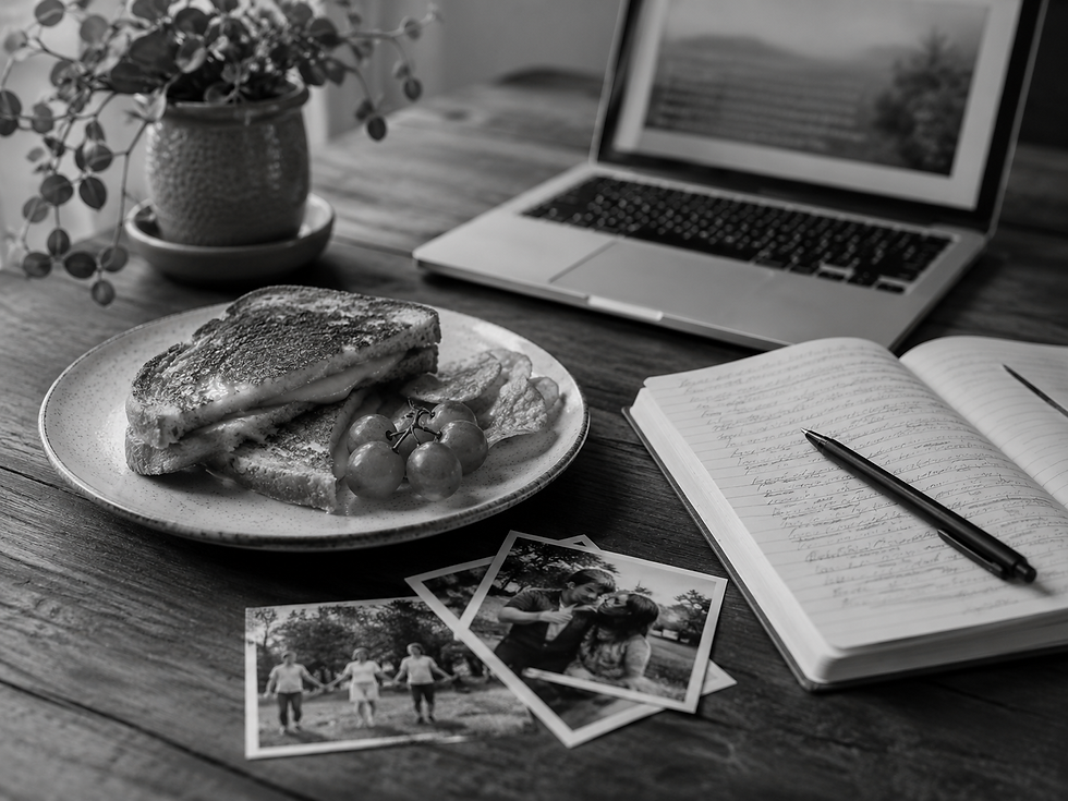 Grayscale desk with grilled cheese sandwich and grapes, open notebook and pen, laptop, plant, and two printed photos.
