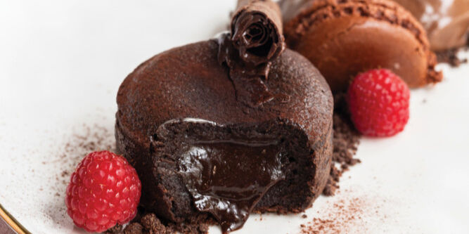 Rich chocolate lava cake with oozing center, garnished with raspberries and chocolate curls, alongside a macaron on a white plate.