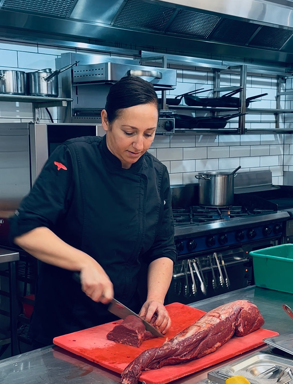 Chef in a black uniform slicing raw meat on a red cutting board in a commercial kitchen with metal pots and pans in the background.