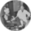 An elderly woman with white hair converses with a man in a lab coat in an office. The woman is seated with her legs crossed, and both appear relaxed.