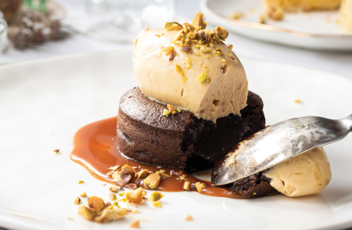 Chocolate fondant with a scoop of caramel ice cream on top, garnished with crushed pistachios. Spoon digging in. White plate, blurred background.