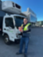 Man in a safety vest holds packaged meat, smiling by a Bidfood delivery truck. Modern building and clear blue sky in the background.