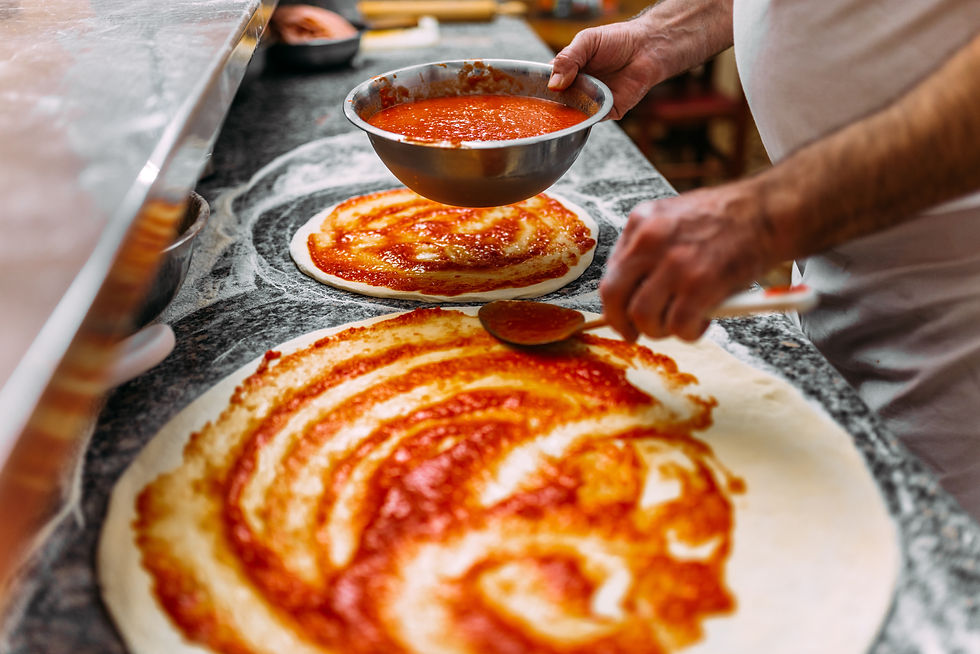 A person spreads red tomato sauce on pizza dough in a kitchen. The dough and sauce contrast against the dark counter. No text visible.
