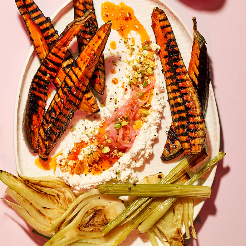 Grilled striped eggplant on a plate with whipped cheese, pickled onions, pistachios, and fennel on a pink background.
