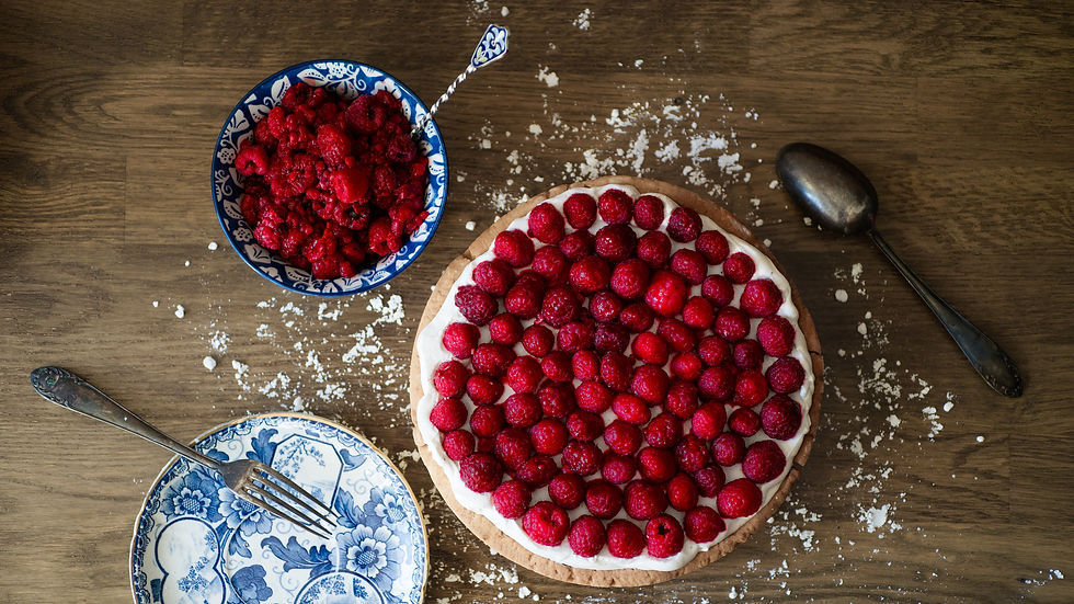 Raspberry tart with a white cream topping on a wooden table, surrounded by a bowl of raspberries, a fork on a floral plate, and a spoon.