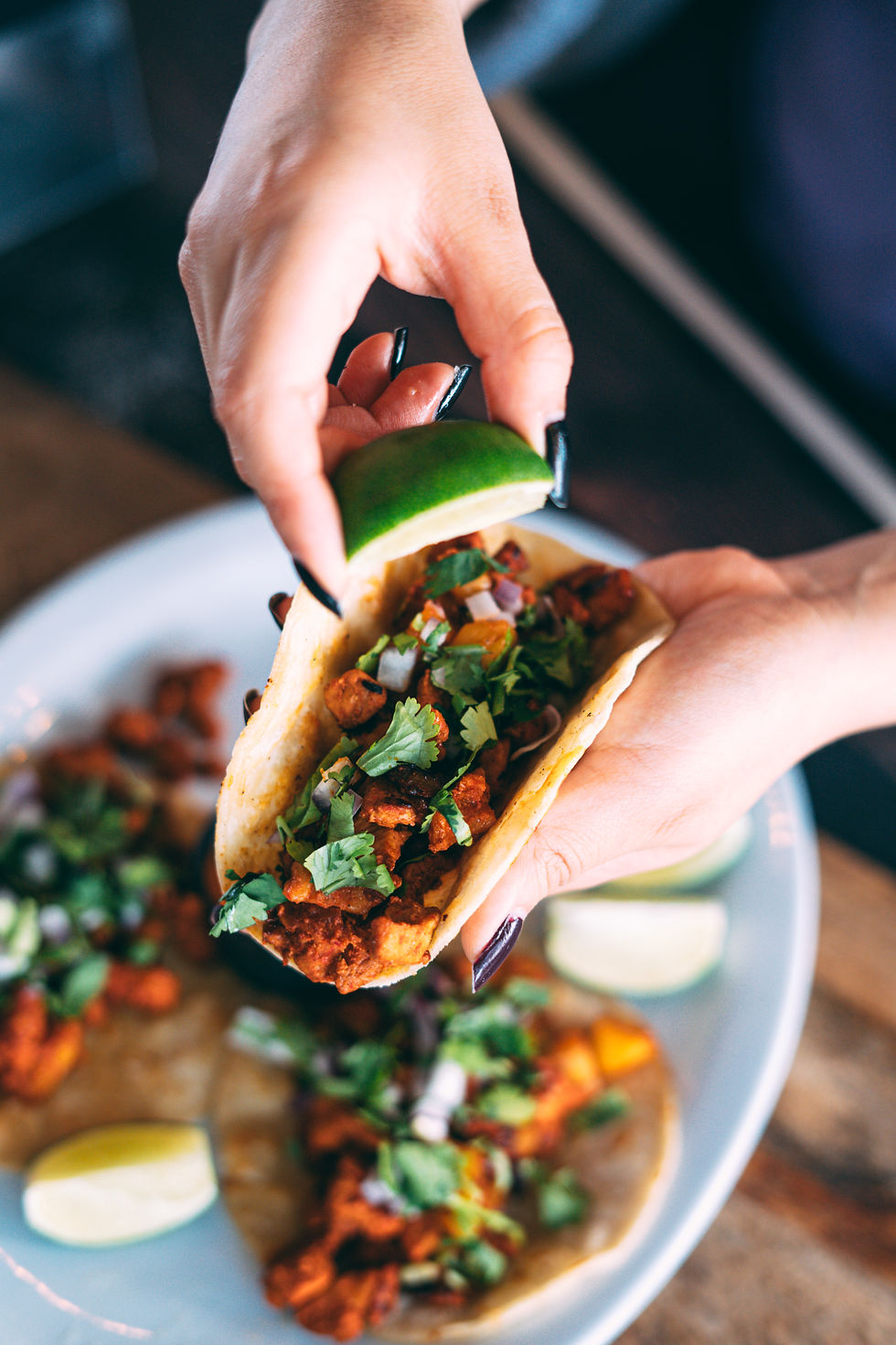 Hands squeeze lime over a taco filled with meat and cilantro. The plate holds more tacos in the background. Vibrant and appetizing scene.
