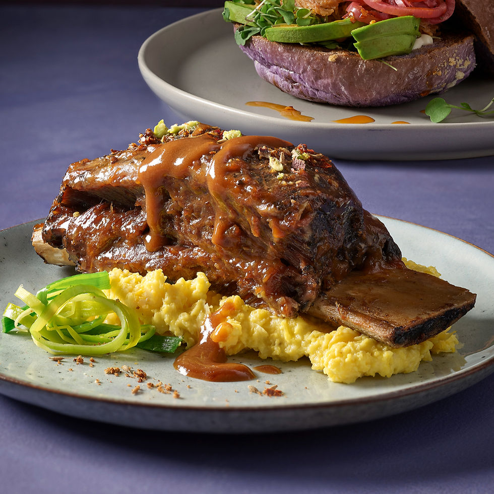 Braised beef rib on yellow mashed potatoes with green garnish on a plate. Background shows a sandwich with avocado on a purple bun.
