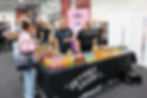 Three people at a drink stand with colorful bottles and a "The Better Drinks Co" banner. A woman with a Bidfood bag converses, smiling.