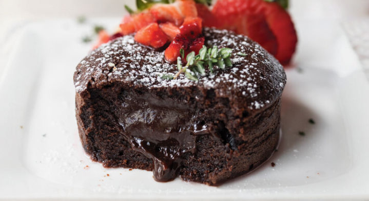 Chocolate lava cake with molten center oozing, topped with strawberries and herbs, dusted with powdered sugar on a white square plate.