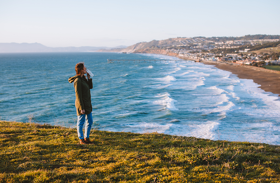 A woman drinking from a bottle of water on the coastline looking out to sea