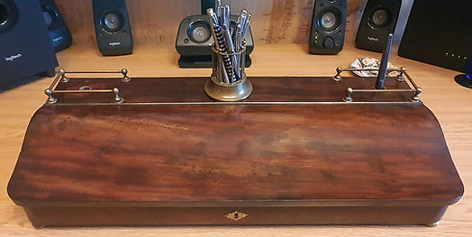 A beautifully reinvented desk storage unit is presented on a wooden workspace, showcasing a complete transformation from its earlier worn condition into a refined, polished, and highly functional piece. The exterior wood has been fully restored, revealing a deep, rich grain with a smooth, satin finish that highlights the natural variations and warmth of the timber.
Across the top, a brass rail system with turned finials adds both structure and elegance, giving the piece a distinctive vintage‑industrial character. Centrally positioned is a brass pen holder filled with assorted pens, further emphasising the piece’s practical use as a desk organiser. The curved front panel has been expertly refinished and fitted with a polished brass keyplate, complementing the overall colour palette and elevating the sense of quality craftsmanship.