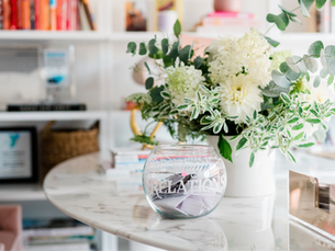 A white vase with lush green and white flowers sits on a marble table beside a glass bowl labeled "RELATIONSHIPS" in a bright, cozy room.