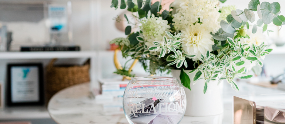 A white vase with lush green and white flowers sits on a marble table beside a glass bowl labeled "RELATIONSHIPS" in a bright, cozy room.