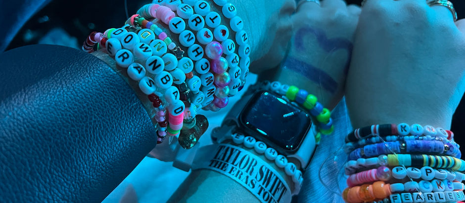 Hands adorned with colorful beaded bracelets, including a Taylor Swift tour wristband, under blue lighting.