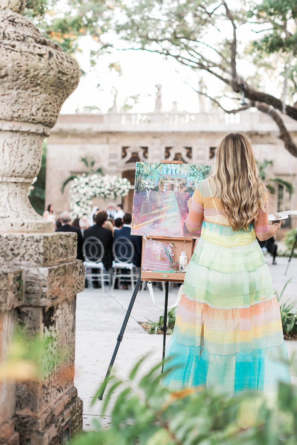 Talia, the artist, in a colorful dress paints outdoors at a wedding. Canvas on easel, stone sculpture, and trees in the background create a serene mood.