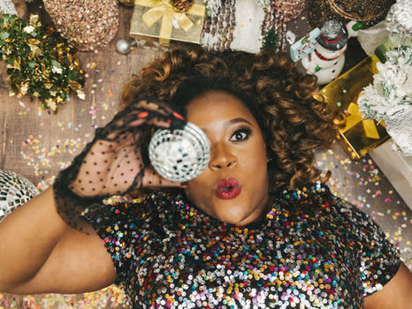 Woman in a colorful sequin top being silly, laying on a confetti-covered floor, covering her eye with a sequin ball