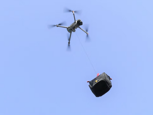 Drone flying against blue sky, carrying a black bag with a thin rope. The bag has small red handles and a yellow tag.