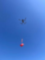 A drone flying in a clear blue sky carries a red and white parachute with a red box. The box has a medical cross and text on it.