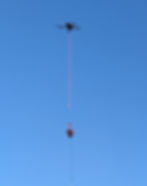 Drone in clear blue sky, carrying a small red basket with a cable. Bright, calm setting with no text visible.