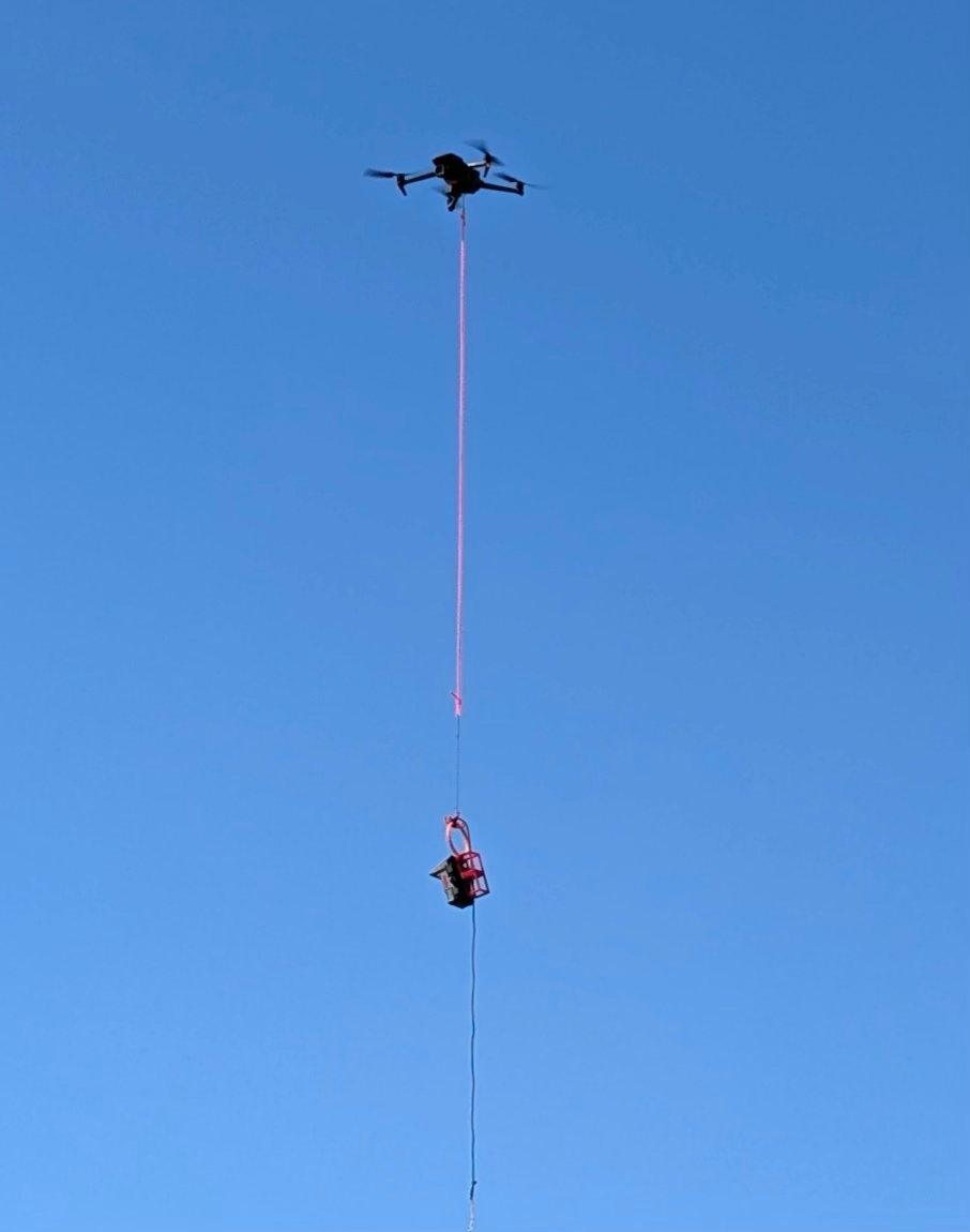 Drone carrying a red birdhouse with a long string against a clear blue sky. The birdhouse dangles in mid-air, creating a whimsical scene.
