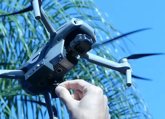 A hand adjusts a gray drone against a clear blue sky, with palm leaves in the background, conveying a tech-focused outdoor setting