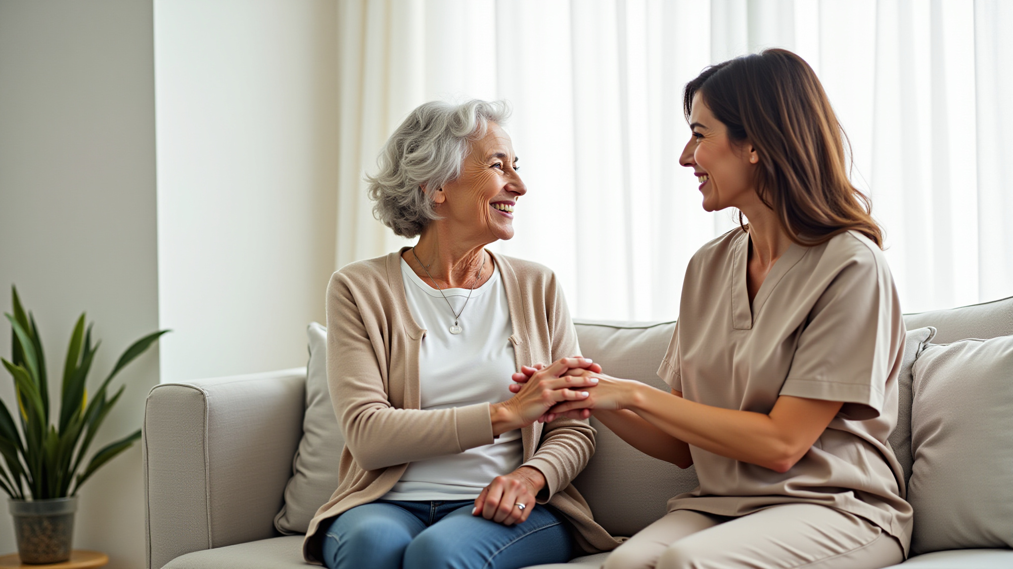 Lachende Pflegerin hält Hand einer älteren Frau auf Sofa.
