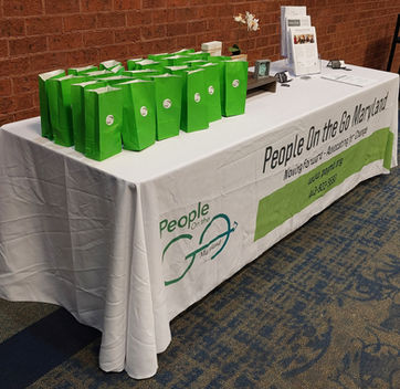 A long rectangular table with a tablecloth printed with the People On the Go logo and information on it holds various meeting materials, including information sheets and welcome bags.