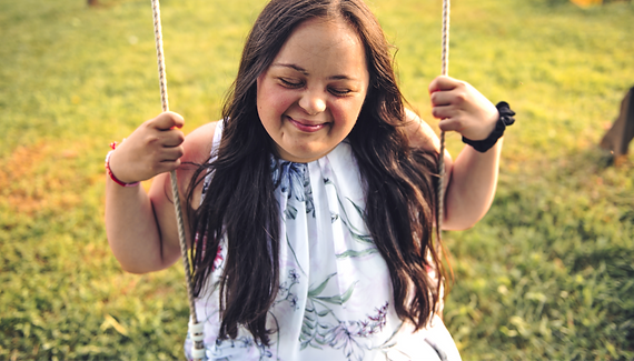 A smiling woman, with intellectual/developmental challenges, swings on a rope swing outside with her eyes closed.