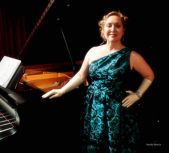 Blonde smiling woman wearing teal one shoulder evening gown, white gloves, and jewelry posing next to black piano.