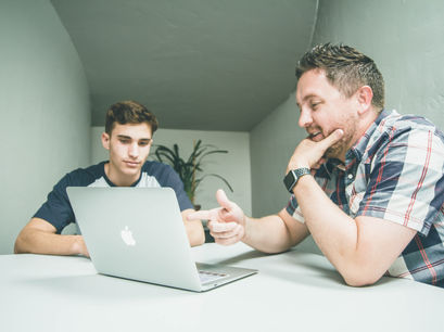 Father and son seated at a table together looking at a laptop while dad provides instructions to his son on screen