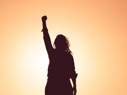 Silhouette of a person with long curly hair illuminated by orange-white lighting, fist raised in the air celebrating a promotion