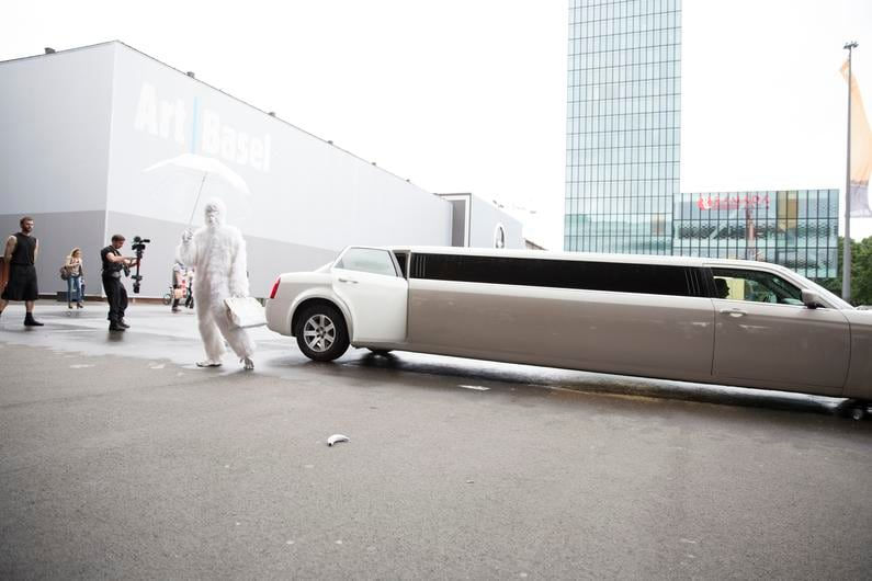 A man dressed head to toe in a white gorilla suit with a white umbrella steps out of a white limousine