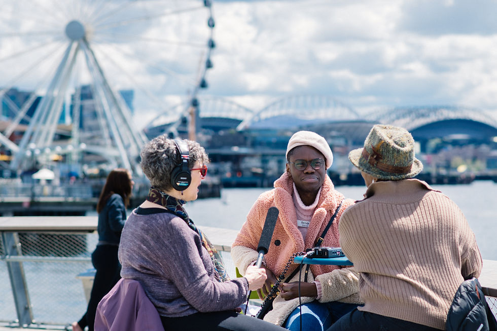 Three people, a woman in a purple shirt with short gray curly hair, a Black male in a pink beret and fuzzy coat, and Black woman in a brown sweater and fedora speak together in from of the Great Wheel (ferris wheel) on Seattle's Pier 62