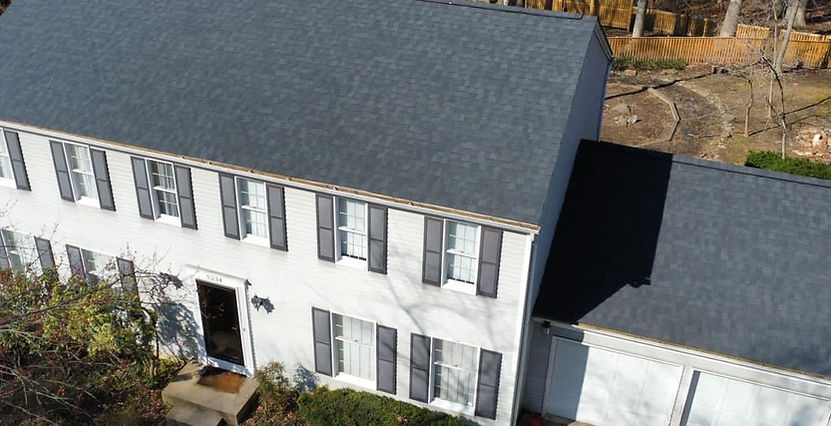Aerial view of two story white house with new dark shingle roof attached garage front door and multiple windows surrounded by trees and backyard fence.