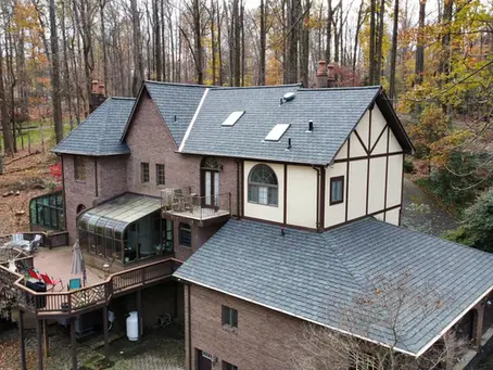 How long does it take to replace a roof? Large brick house with multi level deck glass sunroom and dark shingle roof surrounded by tall trees in autumn.
