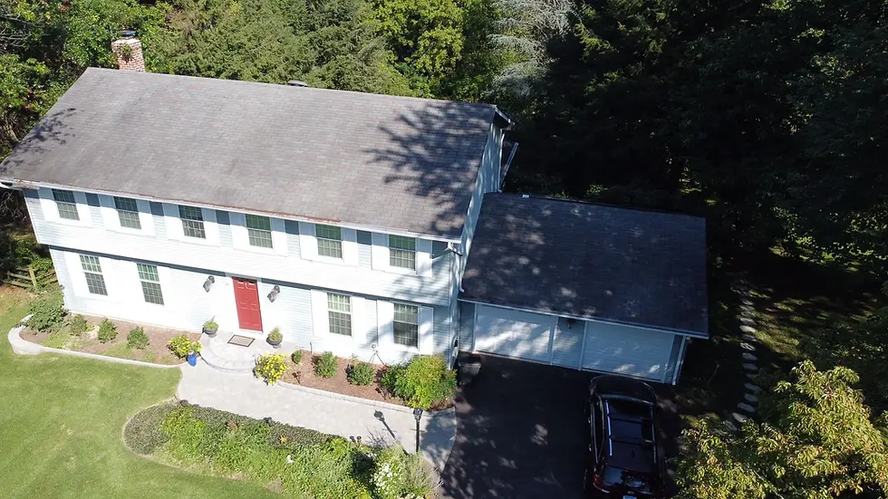Aerial view of two-story white house with red door attached garage dark shingle roof surrounded by trees driveway and landscaped front yard.